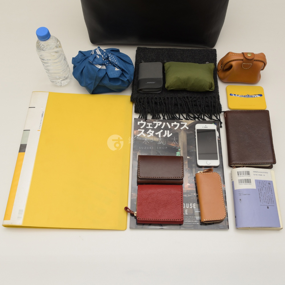 黑色皮革托特包內展示多樣物品，包含一瓶水、藍色印花布包、黑色圍巾、黑色卡夾、軍綠色收納袋、褐色皮革零錢包、黃色卡套、日文書、iPhone手機、深棕色皮革長夾、紅色皮革零錢包、棕色皮革鑰匙包。