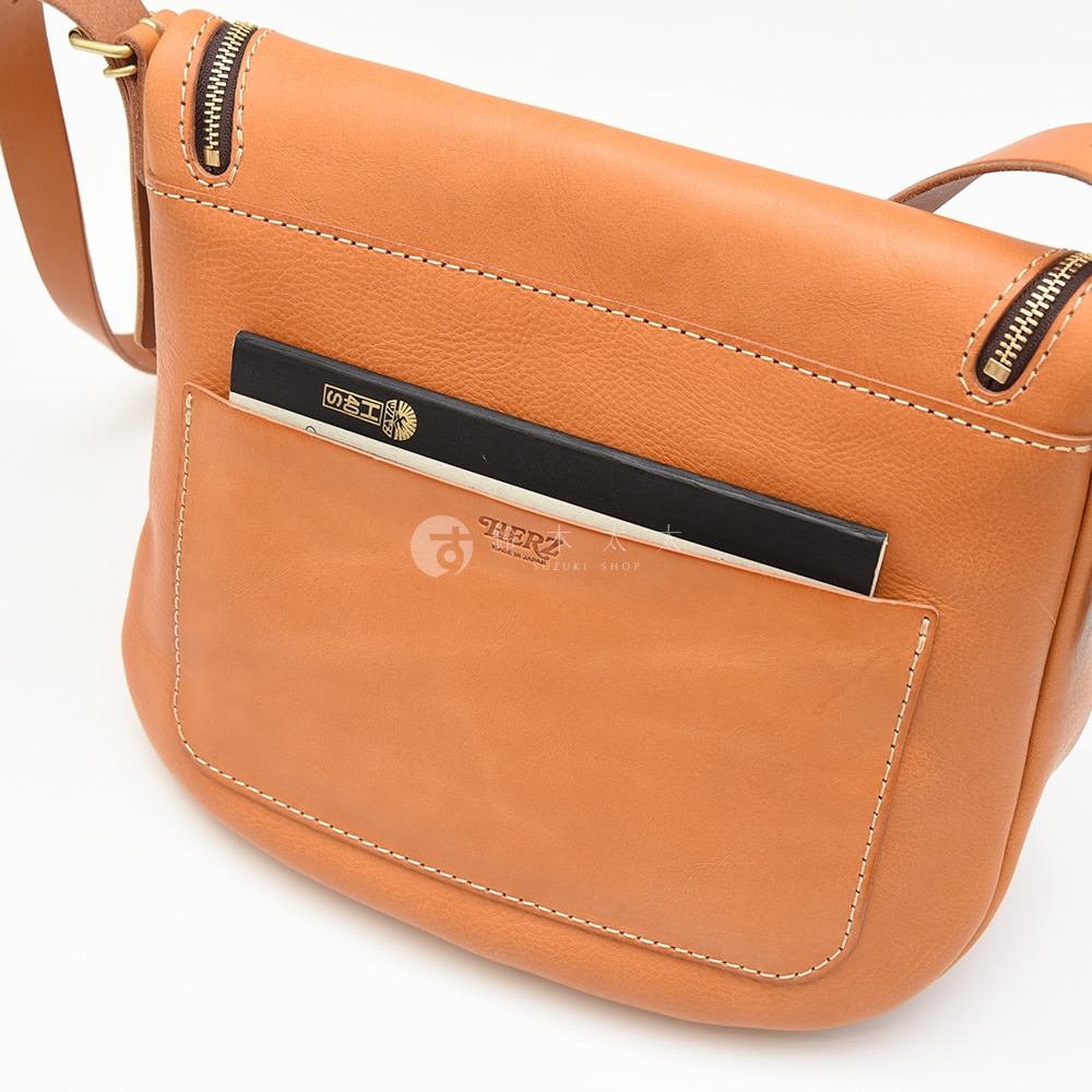 日本HERZ職人手作皮革馬鞍包，特寫正面翻蓋口袋，內含一本黑色筆記本，以及HERZ品牌標誌，整體為棕褐色皮革材質。