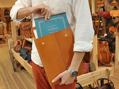 職人手作的棕色皮革信封式雙釦手拿袋，展示其光滑皮革紋理與精緻縫線，袋面有金色釦環，適合收納文件或平板。