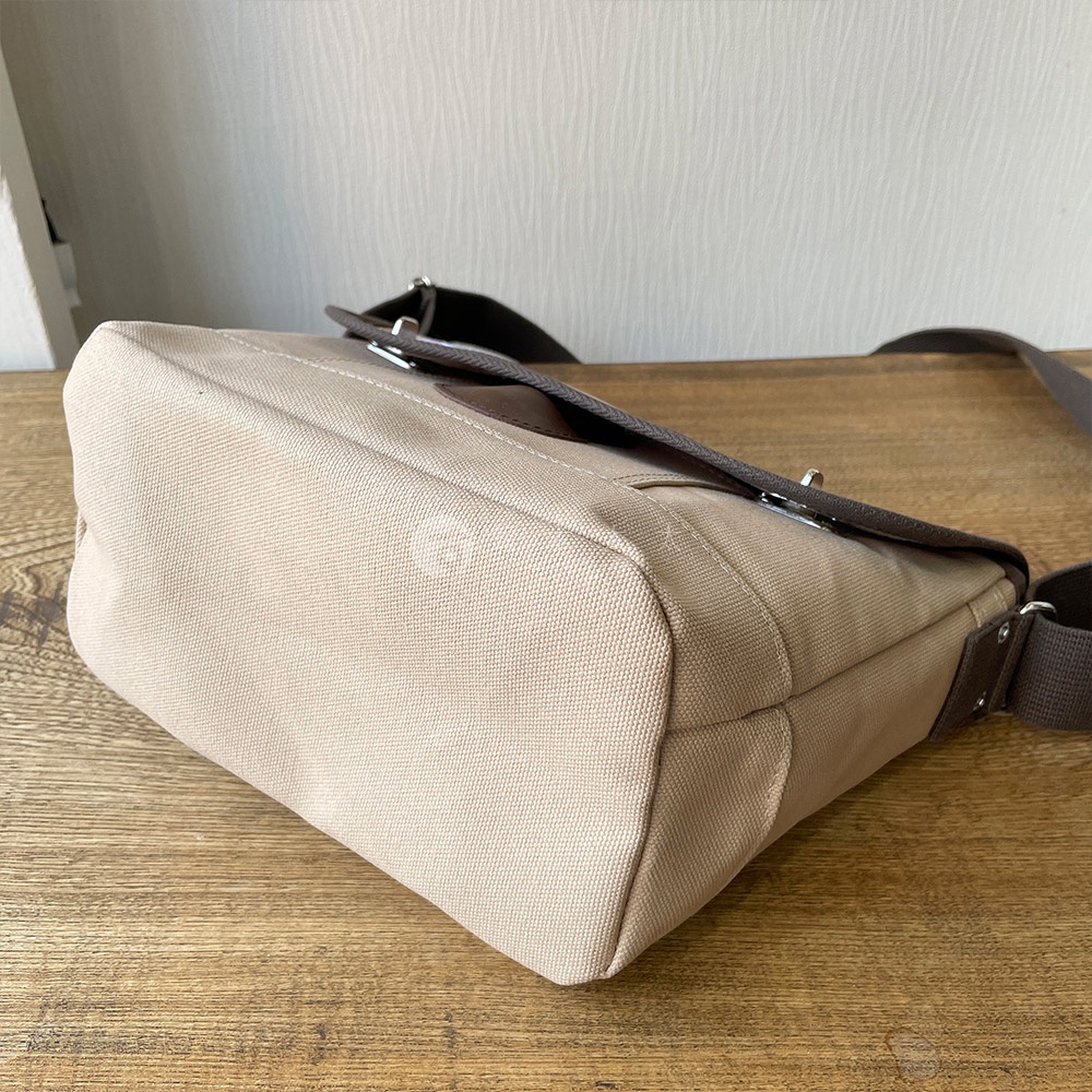 米色純棉帆布雙扣郵差包，搭配深棕色皮革飾邊與肩帶。