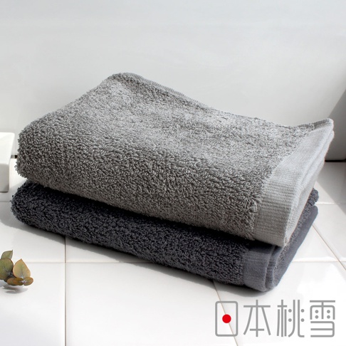 日本桃雪品牌字樣，兩條堆疊的灰色系毛巾，上方為淺灰色，下方為深灰色，置於白色瓷磚背景上，左側有綠色枝葉裝飾。