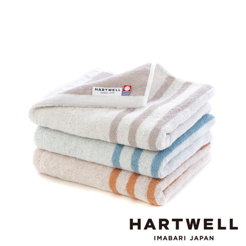 一疊今治毛巾，由上至下分別為米白色、淺藍色、淺粉橘色，皆帶有條紋設計。最上面一條毛巾可見「HARTWELL IMABARI JAPAN」品牌標籤，材質為棉。