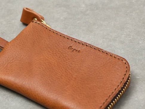 職人手作皮革Organ抽繩收納鑰匙包，採用棕褐色皮革製成，帶有質樸紋理，頂部有抽繩開合設計，並配有金色拉鍊及品牌Logo壓印。