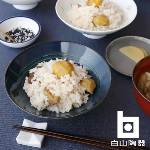 藍色陶製平茶碗盛有栗子飯，碗邊有綠色條紋。旁邊還有白色小碗裝有芝麻，以及灰色小碟盛有醃漬物。