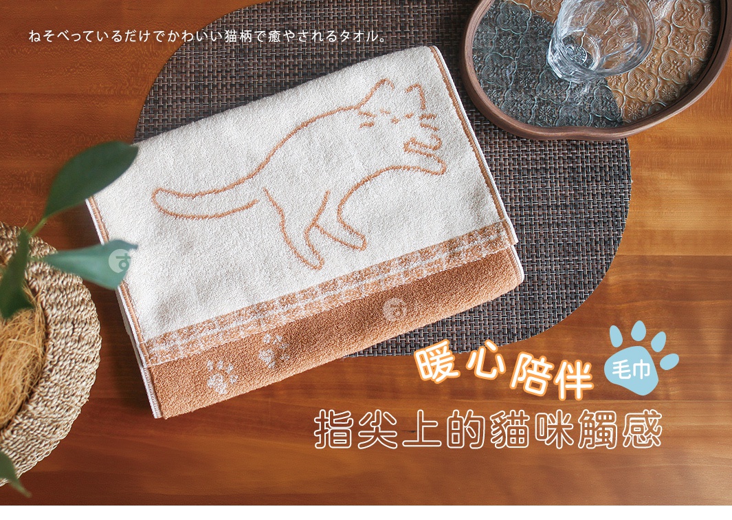 一張俯視視角的照片，展示一條 HARTWELL 今治品牌白色與淺棕色雙色毛巾，毛巾中央繡有可愛的貓咪輪廓，底部綴有細緻的貓爪印花，毛巾整齊擺放在深色圓形編織餐墊上，旁邊還有一盆綠色植物與玻璃杯，整體氛圍溫馨療癒。