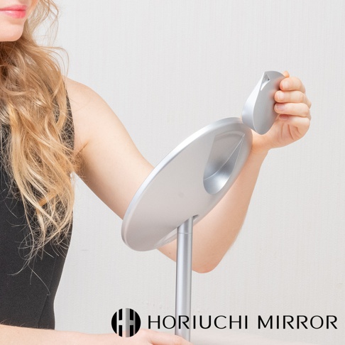 銀色金屬材質的桌上型化妝鏡，圓盤狀鏡面設計，可附加一個方形小鏡，由一名女子操作，背景為白色。