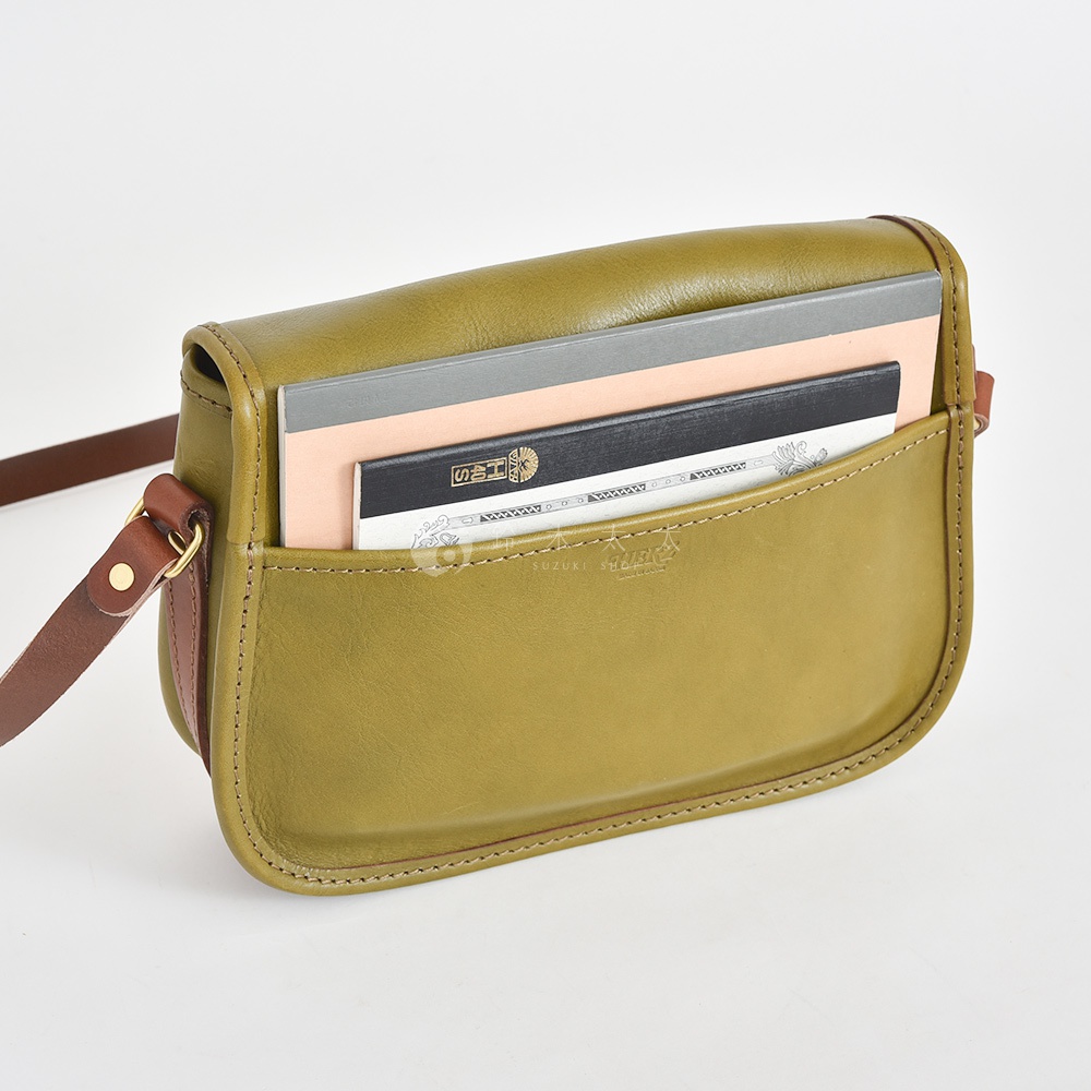 職人手作皮革傳統釣魚包，橄欖綠色，附有深棕色皮革肩帶。包包前方有一個打開的口袋，裡面裝有幾本不同大小的筆記本。