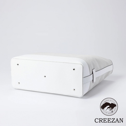 從下方視角拍攝的CREEZAN白色托特包，底座採用細緻紋理皮革，設有五個銀色金屬腳釘以保護包底，連接處以皮革飾帶與帆布包身銜接，整體呈現簡約質感與精緻工藝。