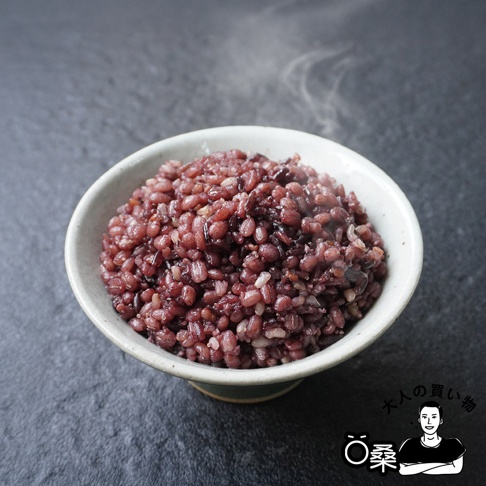 一碗盛滿的日本國產十六穀米，呈現深紫色與紅褐色，米粒飽滿，散發著微微熱氣。碗為淺灰白色，碗底帶有綠色邊緣，置於深灰色背景上。