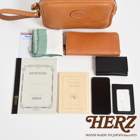 職人手作皮革可拆式扣環萬用手提包，棕色皮革材質，附有可拆卸的腕帶，包包上方有「HERZ」品牌標誌。旁邊擺放著一個棕色長夾、一個黑色皮夾、一本筆記本、一本淺黃色書籍、一支手機和一個卡夾。