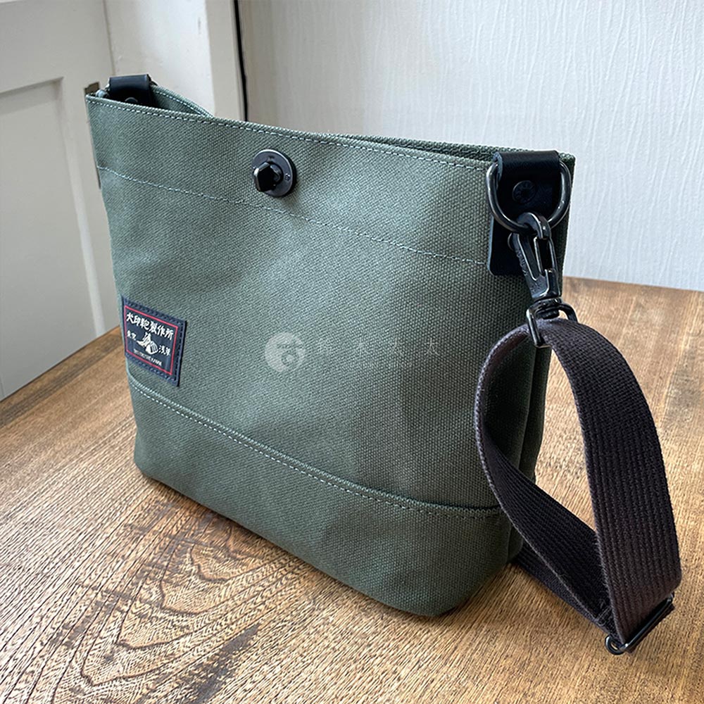 犬印鞄製作所綠色帆布迷你側背包，有黑色揹帶與金屬釦環，正面有紅色標籤。