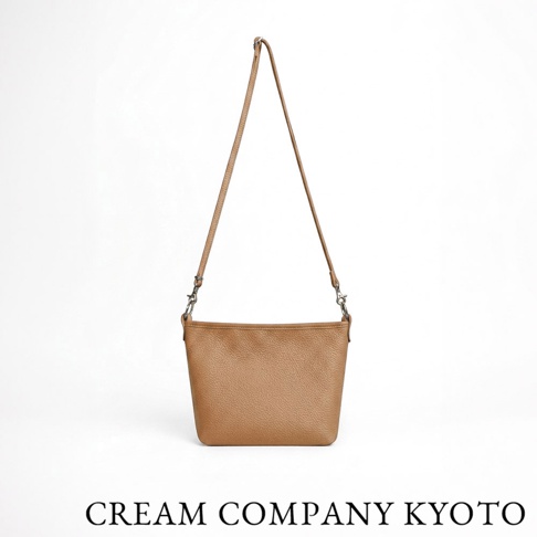 一個棕色皮革材質的 CREAM COMPANY KYOTO 側背包，包身呈現梯形，正面紋理細緻，配有可拆卸的細長肩帶與金屬掛鉤，整體風格簡約輕便。