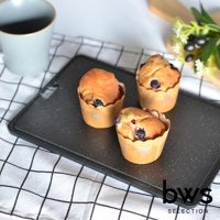 黑色紋理冷卻盤上擺放著三個紙杯裝的藍莓瑪芬，瑪芬烤至金黃色，內含藍莓及少許紅色漿果。盤子左側有一灰色馬克杯，背景為白色，鋪有黑白格紋桌布。