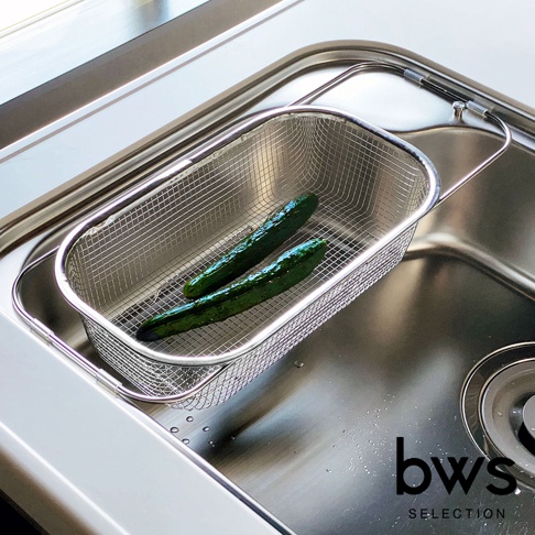 一個不鏽鋼網狀瀝水籃，內有兩條綠色小黃瓜，瀝水籃放置於不鏽鋼水槽中，瀝水籃有可伸縮的邊緣，整體為金屬銀色。