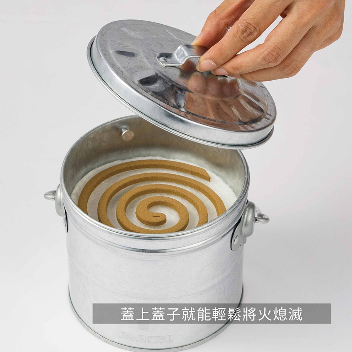 模式B： 一個鍍鋅鋼製的蚊香桶，裡面放著盤狀蚊香，一隻手正準備蓋上金屬蓋子，蓋子上印有中文文字。