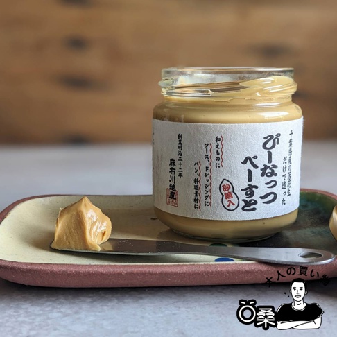 玻璃罐裝的柔滑花生醬，瓶身貼有白色標籤，上面印有日文文字。一小撮金黃色的花生醬置於金屬抹刀上，抹刀放在一個淺色的陶瓷盤子裡。