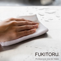 一隻手正使用白色超極細纖維擦拭布擦拭有水滴的銀色檯面，擦拭布質地柔軟，表面有網格紋理。