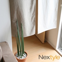 日製手作系輕極簡原色門簾，由米白色及卡其色棉麻布料製成，垂掛於室內空間，一旁擺放著一盆綠色劍蘭植物，種植於陶盆中，盆土覆蓋著白色小石子。