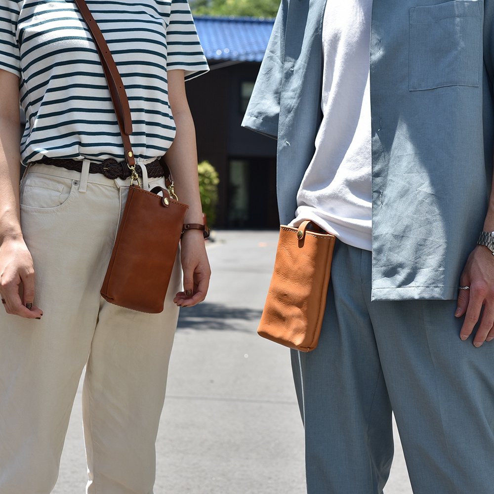 職人手作皮革兩用手機包，展示側背於女性身上及夾於男性褲頭，皆為棕色皮革材質，款式簡約。