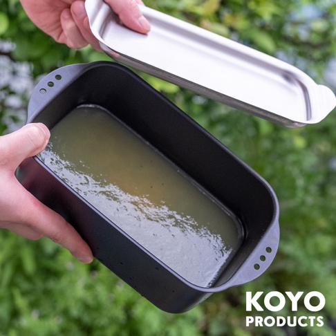 一雙手正舉著 KOYO PRODUCTS 方型鐵製炸物鍋，鍋內裝有液體，上方懸浮著與鍋身配套的不鏽鋼鍋蓋，背景為模糊的戶外綠色植栽。