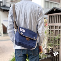 深藍色純棉帆布郵差包，有棕色飾邊和翻蓋，配有兩個金屬扣和一個皮質圓形標籤。包帶為棕色。