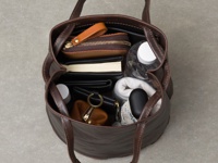 職人手作棕色皮革抽繩肩背包，內有水瓶、筆記本、錢包、化妝包、鑰匙圈等物品，展現包款的收納空間。