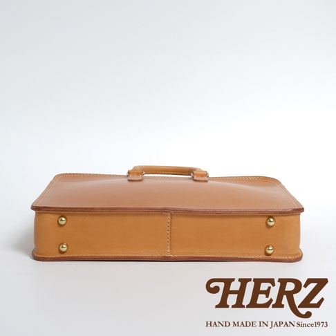 淺棕色皮革手提商務公事包，配有提把，包身側面有金屬釘裝飾，縫線清晰，風格簡約。