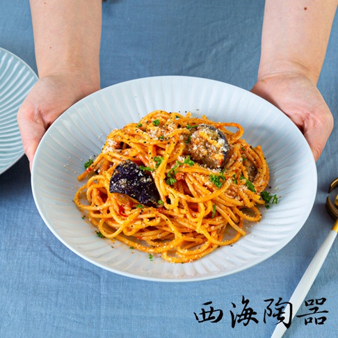 淺藍色波佐見燒 FUWARI 主食盤裝盛著茄子番茄義大利麵，麵條裹滿醬汁，撒有起司粉與香草碎，盤邊有扇形壓紋，雙手捧著盤子。