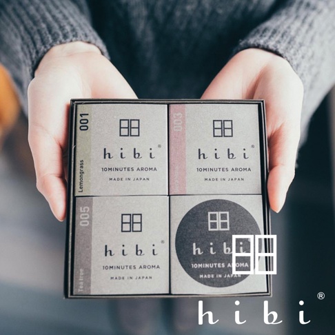 雙手捧著一盒方形禮盒，盒內裝有四個印有 hibi 品牌標誌的灰色小方盒，分別標示著 001 檸檬草、003 天竺葵、005 茶樹等不同香氛款式的包裝，整體呈現出簡約精緻的日系質感。