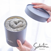 一條灰色、蓬鬆柔軟的浴巾捲成圓形，置於灰色圓筒形包裝盒內，盒蓋由一隻手拿起，另一隻手托著浴巾。