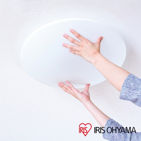 一隻手觸碰著白色圓盤狀的吸頂燈，另一隻手輔助固定，燈體為平坦的圓形設計，表面光滑，背景為白色紋理牆壁。