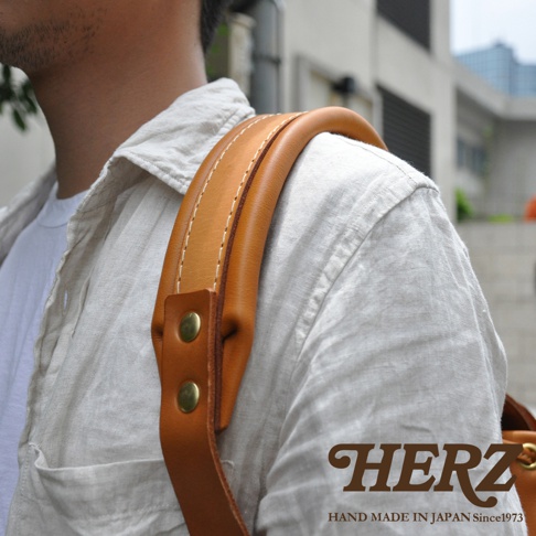 職人手作皮革復古風旅行後背包，展示其棕褐色皮革肩帶，帶有縫線和兩顆黃銅鉚釘，搭配淺色亞麻襯衫。