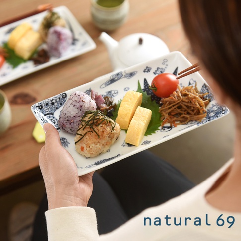 一只手拿著一個白色的長方陶瓷餐盤，盤內盛有日式早餐，包括兩塊玉子燒、兩顆飯糰、番茄、醃漬物及牛蒡絲，餐盤邊緣飾有藍色魚形和海浪圖案。