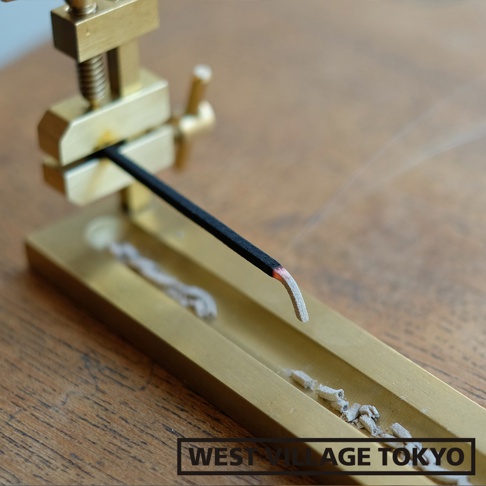 黃銅夾式水平線香架，夾持一根燃燒中的黑色線香，線香末端呈現紅熱狀態，並有細微的煙霧飄出。線香架為金屬材質，表面光滑，呈現黃銅色澤，底部有收集香灰的槽狀設計。