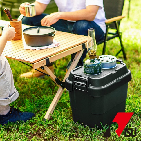 黑色塑膠可堆疊收納箱，箱蓋上放有露營燈和瓦斯罐，置於戶外草地上。