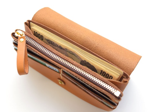 職人手作皮革旅行用長夾，淺棕色皮革材質，內含多疊日圓鈔票與信用卡，拉鍊及金屬扣環為銀色。