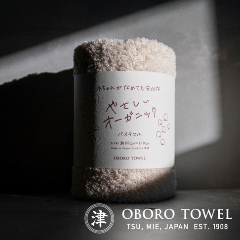 捲起的米白色有機棉浴巾，質感柔軟，上面印有日文「やさしいオーガニック」及「OBORO TOWEL」品牌字樣，標示尺寸約60x120公分，日本製造，100%純棉。
