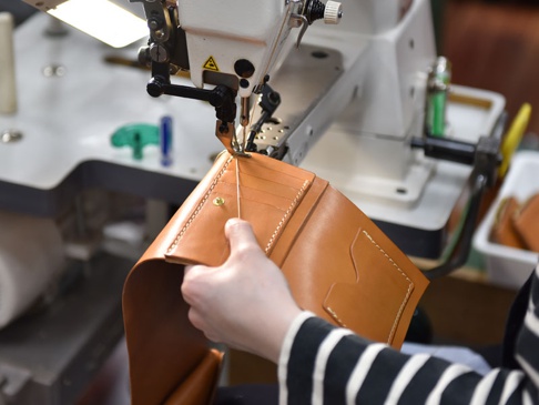職人正在使用縫紉機製作一個棕色皮革中型雙折短夾，可見精緻的縫線與卡槽。