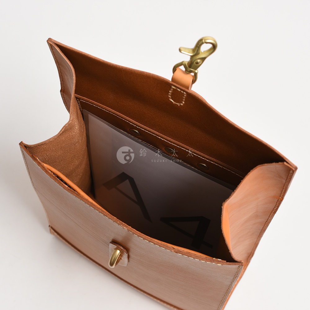 職人手作皮革紙袋風後背包，棕褐色皮革材質，內部可見A4文件夾，包款頂部有黃銅色鉤扣。