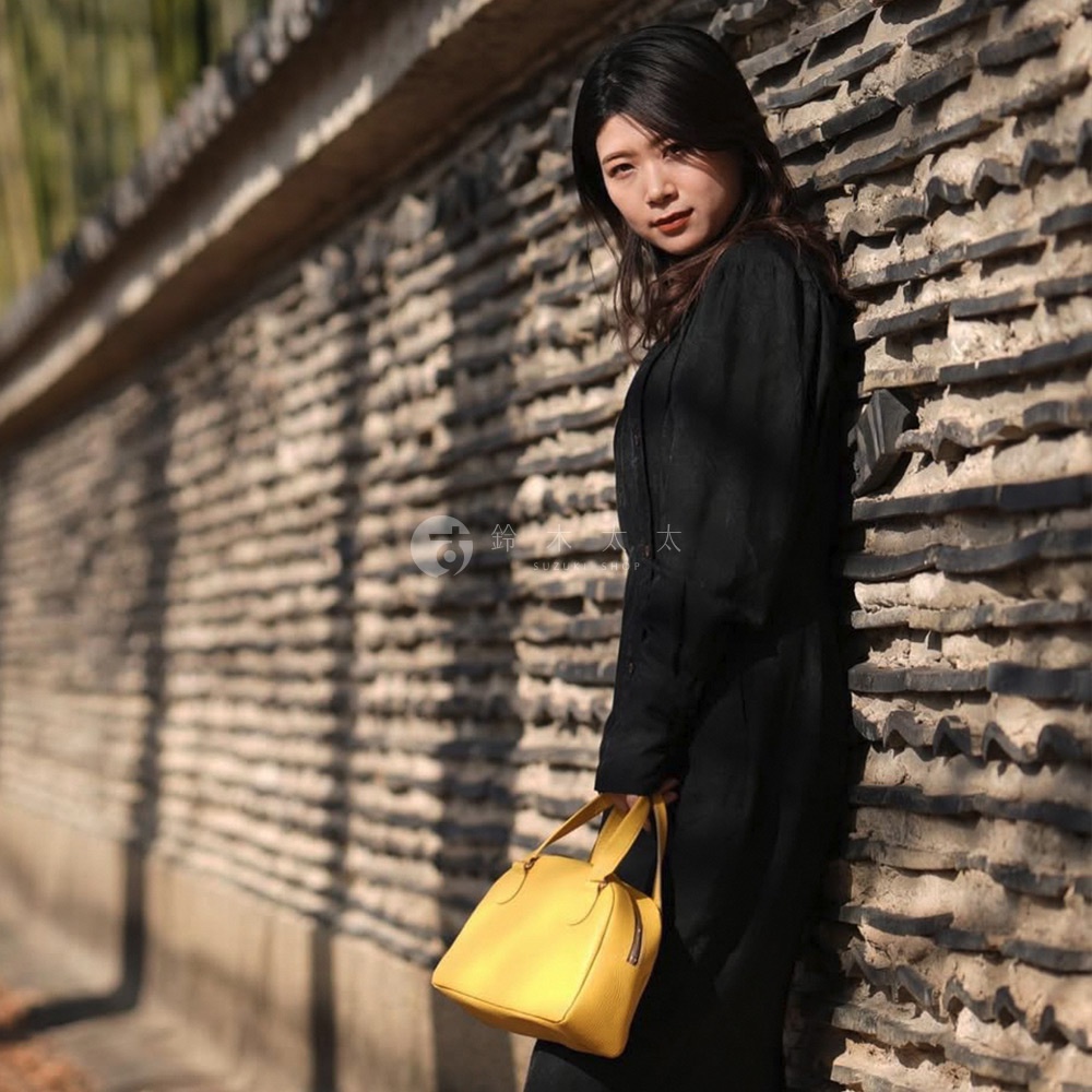 手持亮黃色半月形迷你手提包，包身皮革紋理清晰，配有雙提把，搭配黑色長袖服裝，背景為斑駁的磚牆。