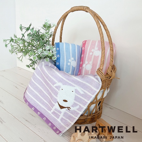 這是一個裝在藤編提籃裡的 Hartwell 日本今治產純棉紗布浴巾，籃中擺放了粉紅色與藍色的條紋浴巾，籃外則垂掛著一條印有白色小狗圖案的紫色條紋紗布浴巾，旁邊配有綠色植物裝飾與木製玩具車，整體呈現簡約溫馨的居家生活感。