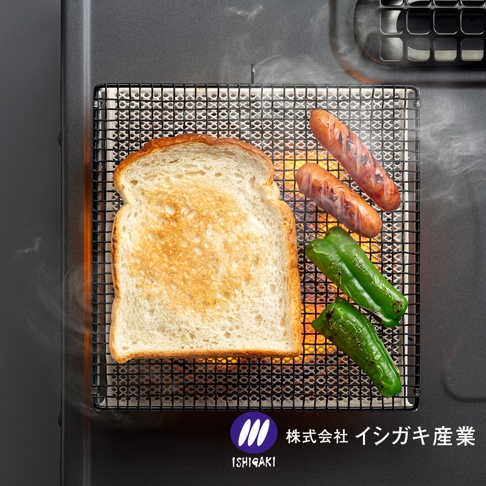 上方視角的照片中，在黑色的瓦斯爐面上放著石垣產業的方形陶瓷烤網，網架上正烤著一片吐司、兩條烤香腸以及兩條青椒，網下透出紅亮的爐火，並有少許白煙飄散，右下角標示著株式会社イシガキ産業的企業標誌。