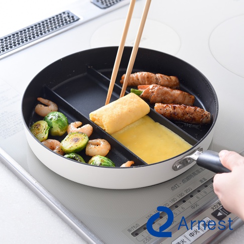 一隻手握著Arnest三合一分格平底鍋的把手，鍋內分隔設計讓蝦子、抱子甘藍、玉子燒與肉捲能同時在鍋中烹調，上方使用筷子翻轉玉子燒，鍋具下方印有電磁爐操作介面。