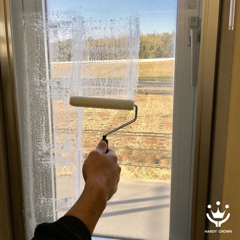 一只手持著帶有米白色滾筒的紗窗清潔刷，正在清潔布滿泡沫的紗窗。紗窗後方可見室外風光。