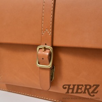 職人手作皮革三用單扣商務後背包 BC-4。近拍特寫，展示部分包款的卡其色皮革主體、一個仿舊黃銅色單扣及皮革繫帶，以及包款底部的手工縫線與品牌標誌。