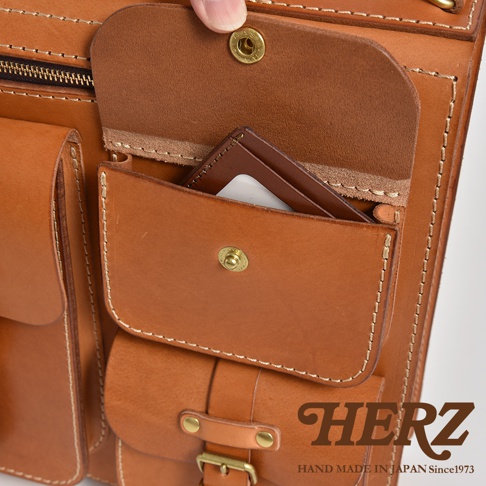 職人手作皮革三口袋三用後背包 A-19-R，特寫近拍，展示多個棕色皮革口袋、金屬鈕扣及縫線細節，左側有拉鍊，右下角為品牌標誌。