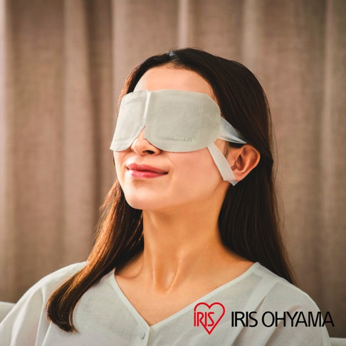 一位長髮女性戴著淺灰色的 IRIS OHYAMA moiscle 蒸氣眼罩，她面帶微笑、頭部微微仰起，背景為柔和的垂簾，整體畫面傳遞出放鬆與舒適的氛圍。
