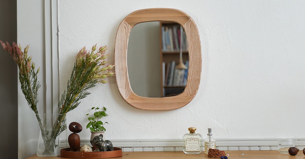 橢圓形木框壁掛鏡，天然木色，鏡面清晰，框邊帶有自然紋理，掛於白色紋理牆面上，左側為玻璃花瓶內插著粉色乾燥花束，右側桌面擺放著香水瓶及裝飾品。