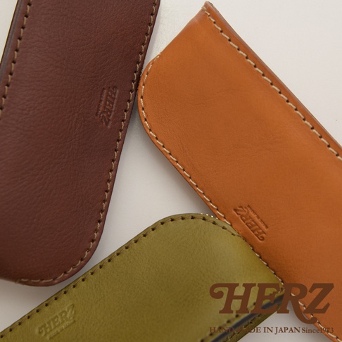 職人手作皮革三角弧形收納筆袋，展示棕色、橘棕色及橄欖綠三款顏色，皆為真皮材質，邊緣以米色縫線手工縫製，皮革表面壓印有「HERZ」品牌標誌。