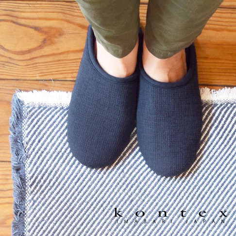 一雙深藍色棉麻室內拖鞋，鞋面有細小的格紋紋理，腳上穿著卡其色褲子，踩在藍白條紋的毛巾地毯上。